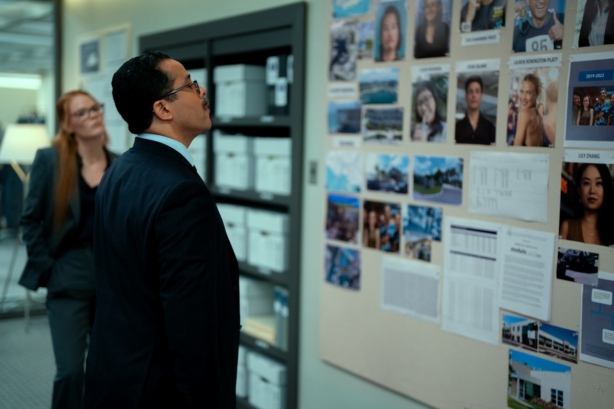 Two people in business attire stand in an office, examining a bulletin board covered with photos, documents, and notes, suggesting an investigation or case analysis setting.