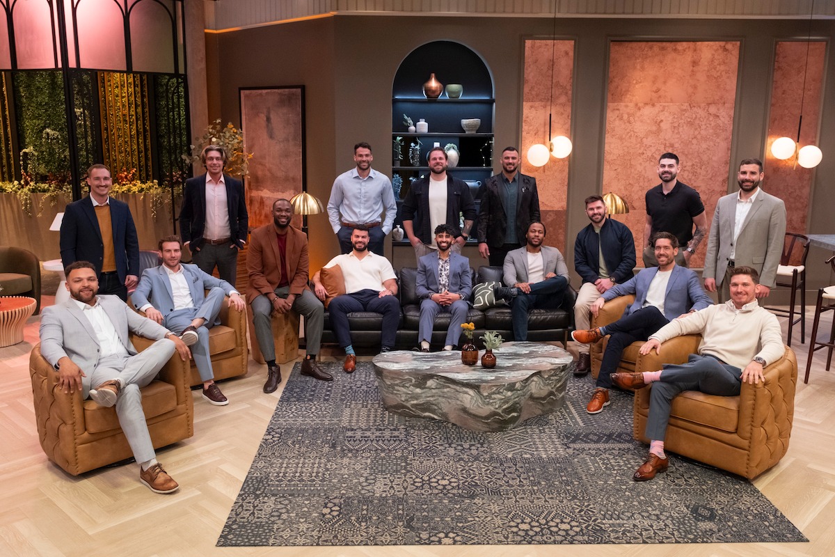 A group of men in suits and smart casual attire pose together in a stylish, modern lounge with armchairs, a large rug, decorative lighting, and shelves with vases in the background.