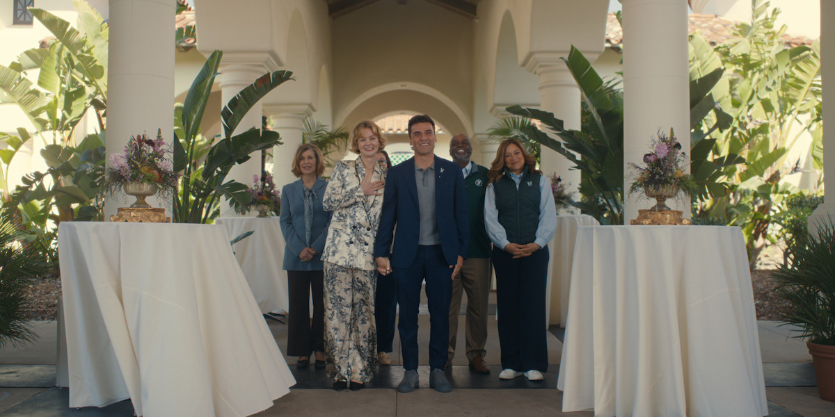 Five people dressed formally stand between columns at an outdoor venue with white tablecloth-covered tables, floral arrangements, and lush green plants.