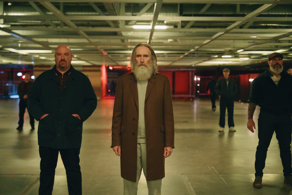 A group of men, including an older man with a long white beard in a brown coat, stand in a dimly lit, industrial-style room with exposed ceiling beams and concrete floors.