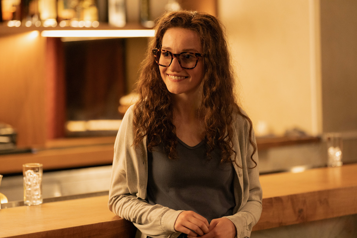 A woman with curly hair and glasses smiles while standing at a wooden bar in a warmly lit, modern indoor setting, with glasses and bottles in the background.
