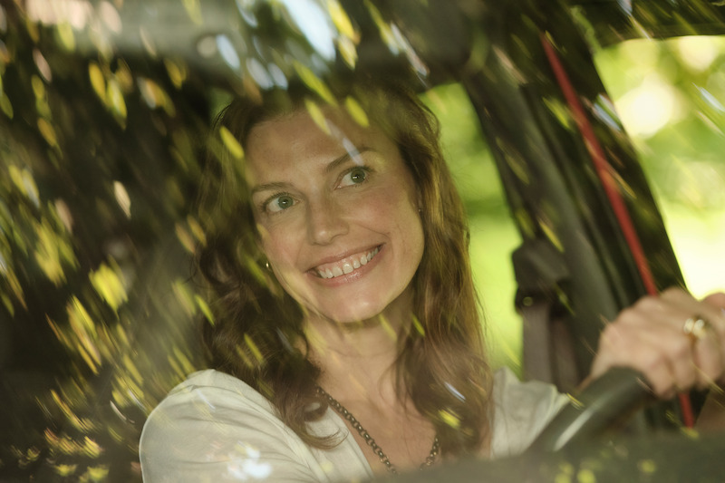 Smiling woman sitting in driver’s seat of a car, seen through windshield with green leafy reflections, natural outdoor lighting, relaxed atmosphere.