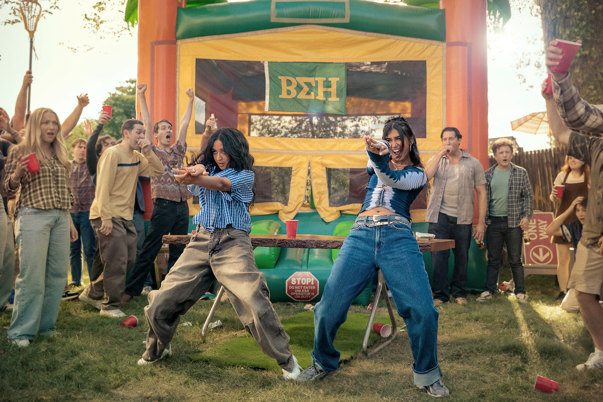 Two women dancing energetically outside at a lively party with a group of people cheering around them, red cups in hand, and an inflatable bounce house with Greek letters in the background on a sunny day.