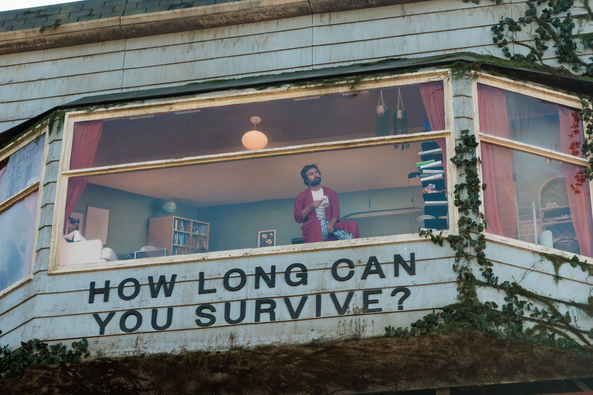 A man stands in the window of a weathered, overgrown billboard, holding a mug inside a cluttered living room. Below the window, a large sign reads "HOW LONG CAN YOU SURVIVE?" with ivy climbing the wall.
