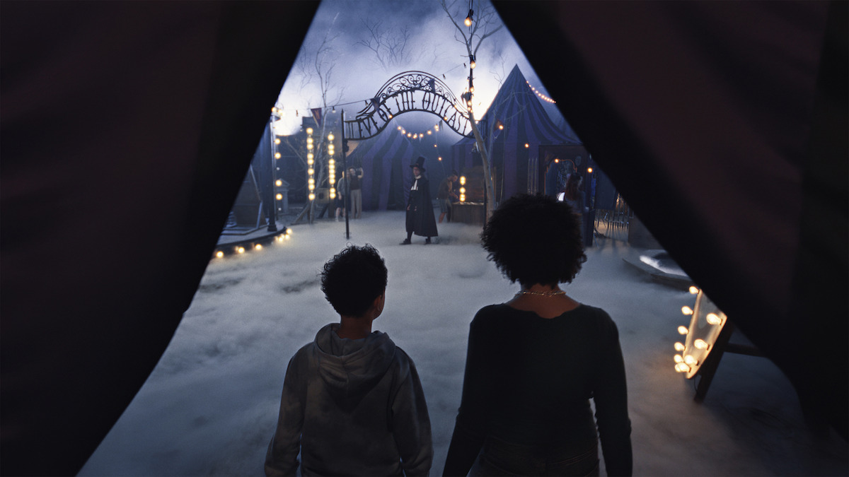 Two people enter a foggy, mysterious carnival at night, with glowing lights, tents, and a figure standing in the center. The scene feels eerie and magical, suggesting anticipation and curiosity.