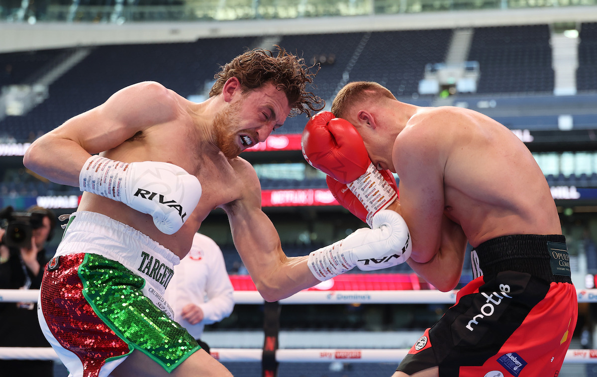 Two male boxers in a boxing ring during a match, one landing a punch on the other who is blocking, with arena seating and spectators in the background.