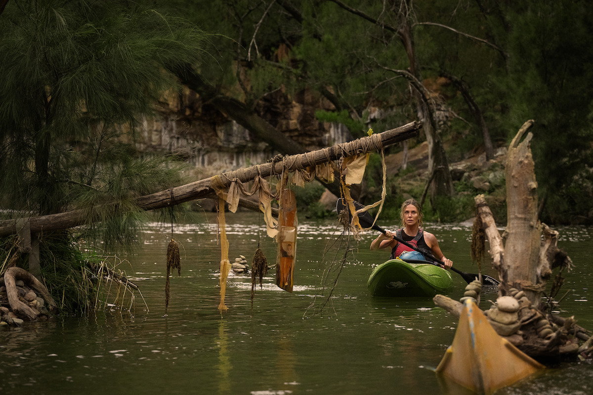 Person kayaking on a river surrounded by trees, approaching a log decorated with fabrics and items hanging down, with a rocky, forested background and debris in the water.