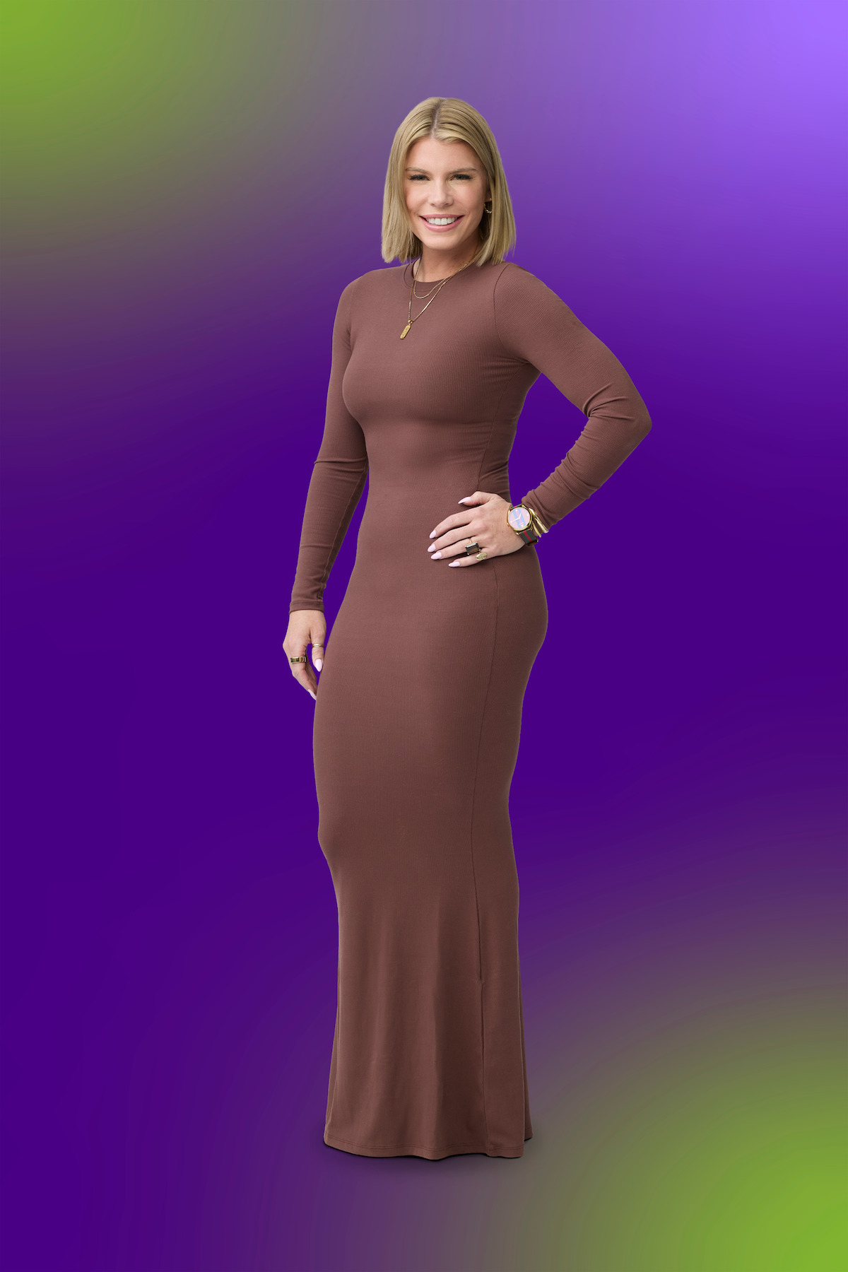 Woman standing and smiling, wearing a long brown dress with long sleeves, gold watch, and necklace, posing confidently against a colorful, gradient purple and green background.