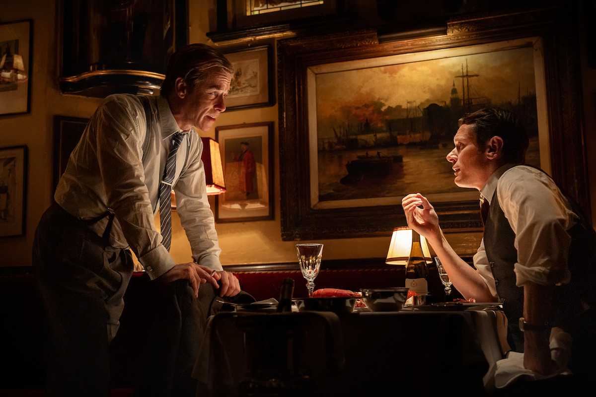 Two men in dress shirts talk at a dimly-lit restaurant table surrounded by framed paintings and warm lighting, creating an intimate, vintage atmosphere.