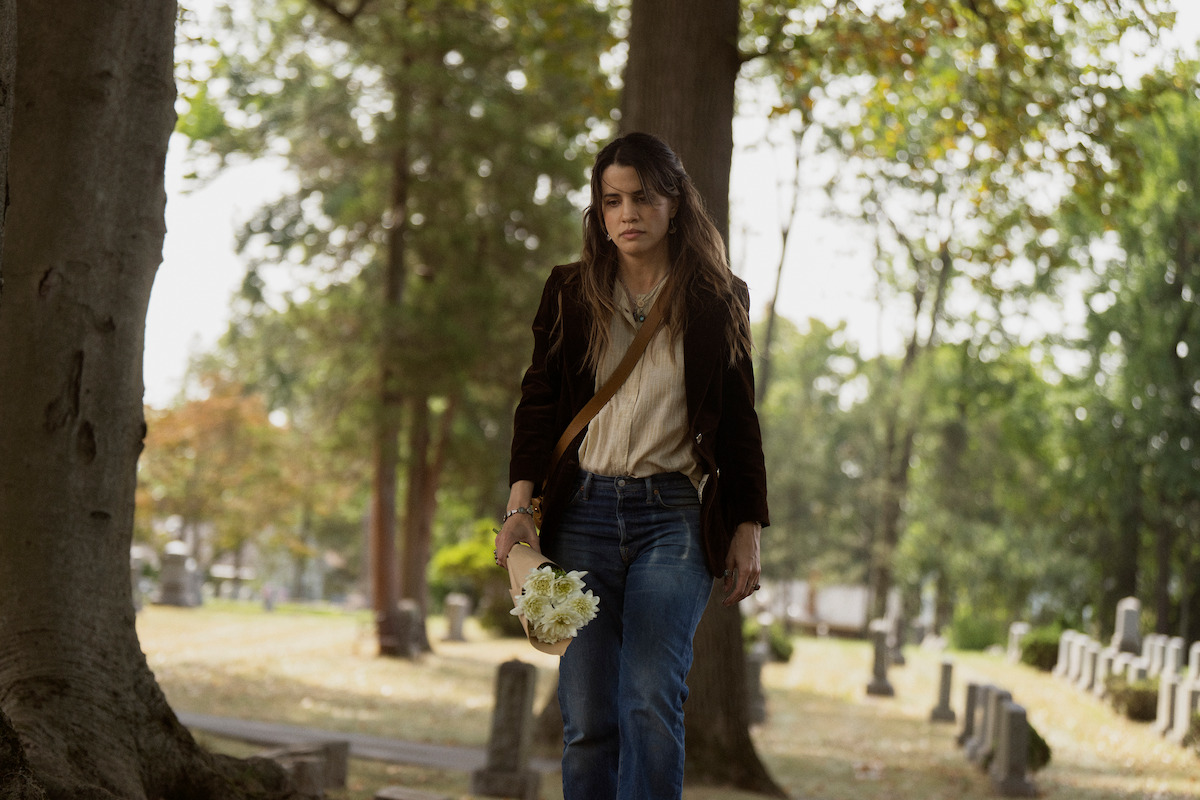 A woman walks solemnly through a cemetery holding white flowers in daylight, surrounded by gravestones and trees, conveying a somber, reflective mood.
