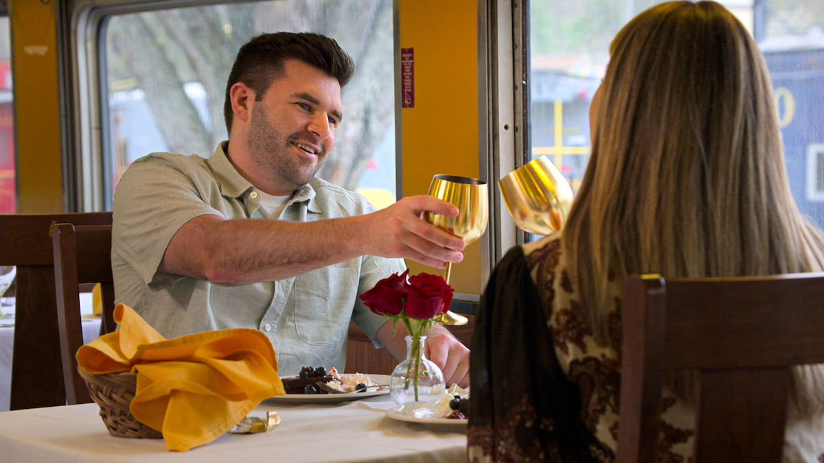 Connor and Brianna toasting with gold cups at a restaurant table set with roses and a bread basket, by a window with trees and yellow walls visible, creating a cozy, romantic dining setting.