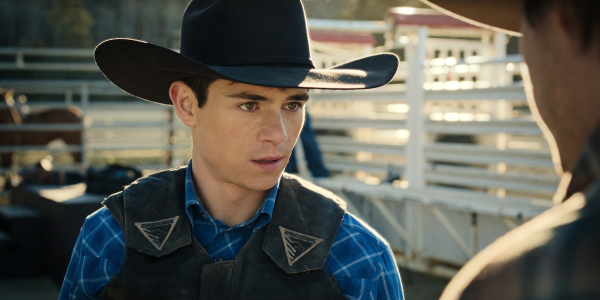 Young man in cowboy hat and plaid shirt stands outdoors at a rodeo or ranch setting, wearing a protective vest, talking to another cowboy with livestock and fencing in the background.