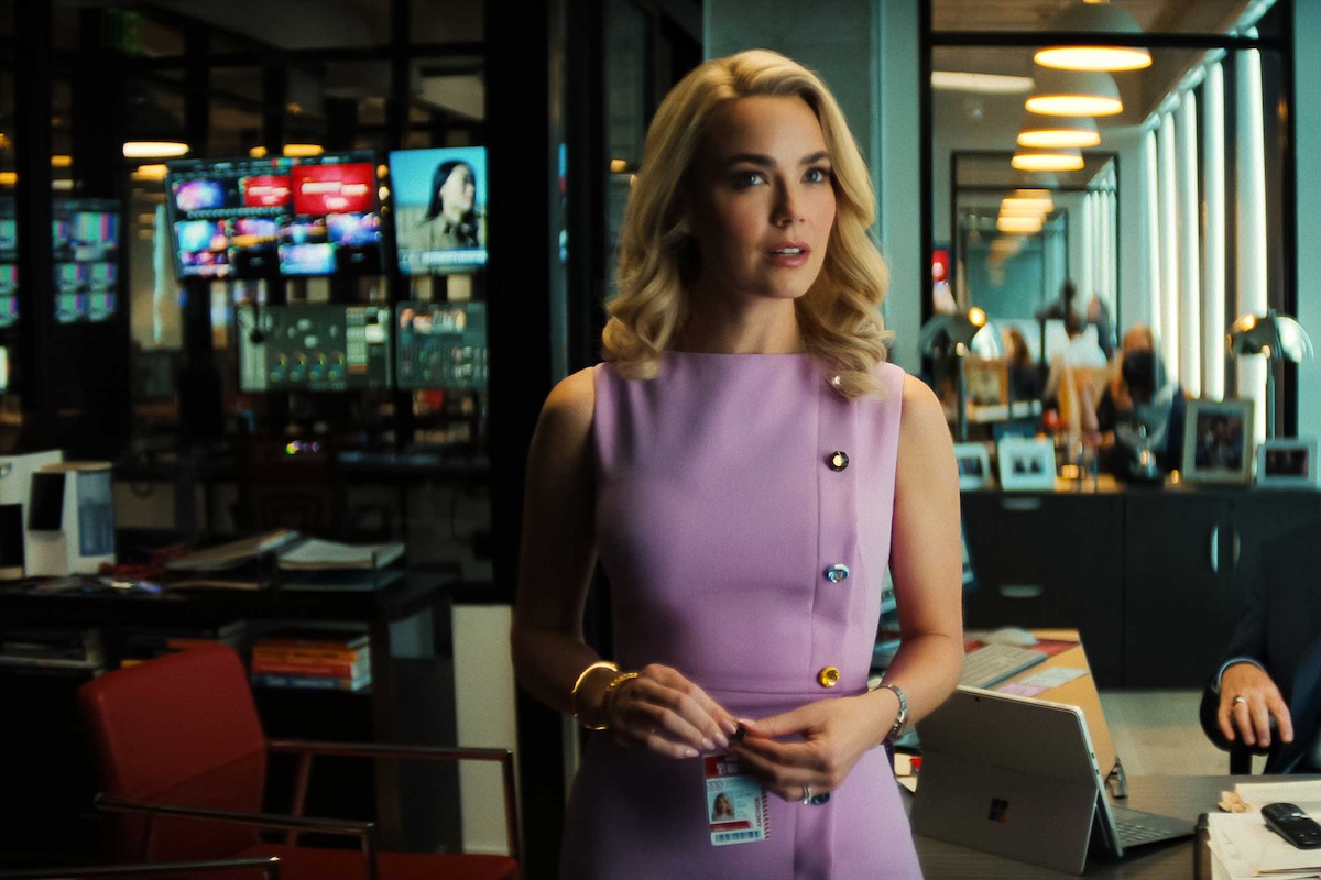 A woman in a lavender dress stands in a modern office with glass walls, bright lighting, red chairs, monitors displaying news, and people working at desks in the background.