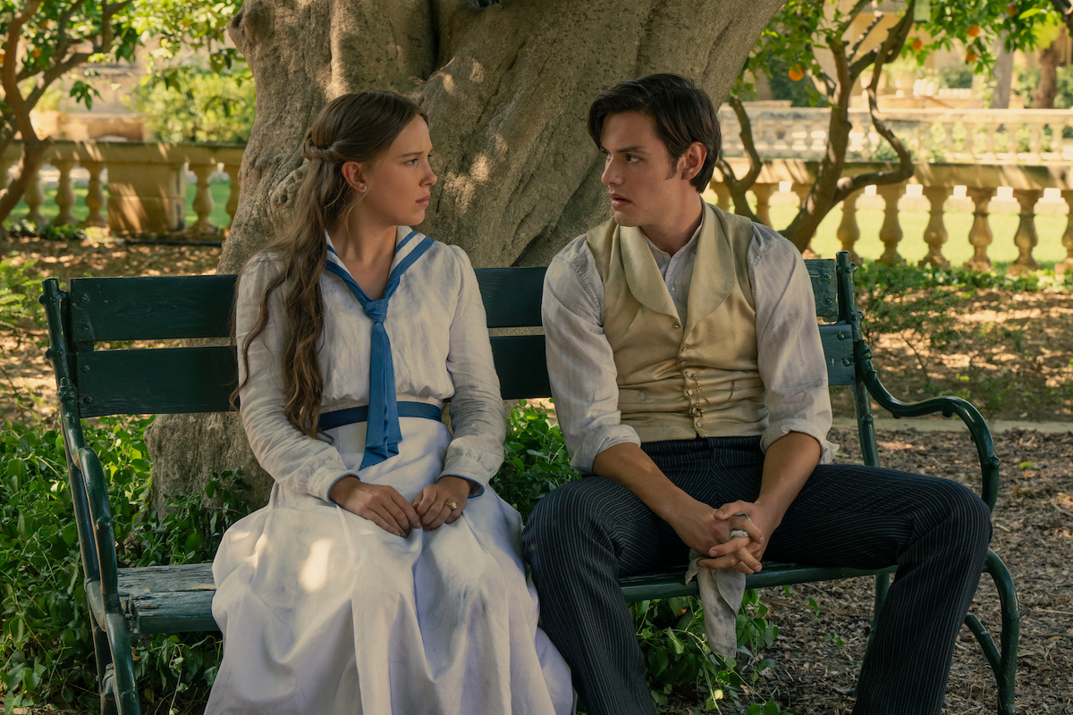 A young woman in a white dress and a young man in a vest and shirt sit on a green bench under a large tree in a sunlit garden, looking at each other with serious expressions.