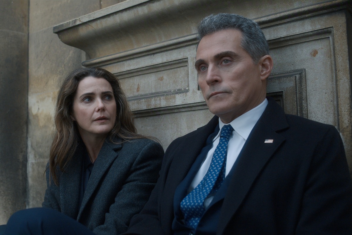 Two people in formal coats sit on outdoor stone steps, appearing serious and deep in conversation, with an old stone wall in the background. The man wears a suit and tie with an American flag pin, suggesting a political or official context.