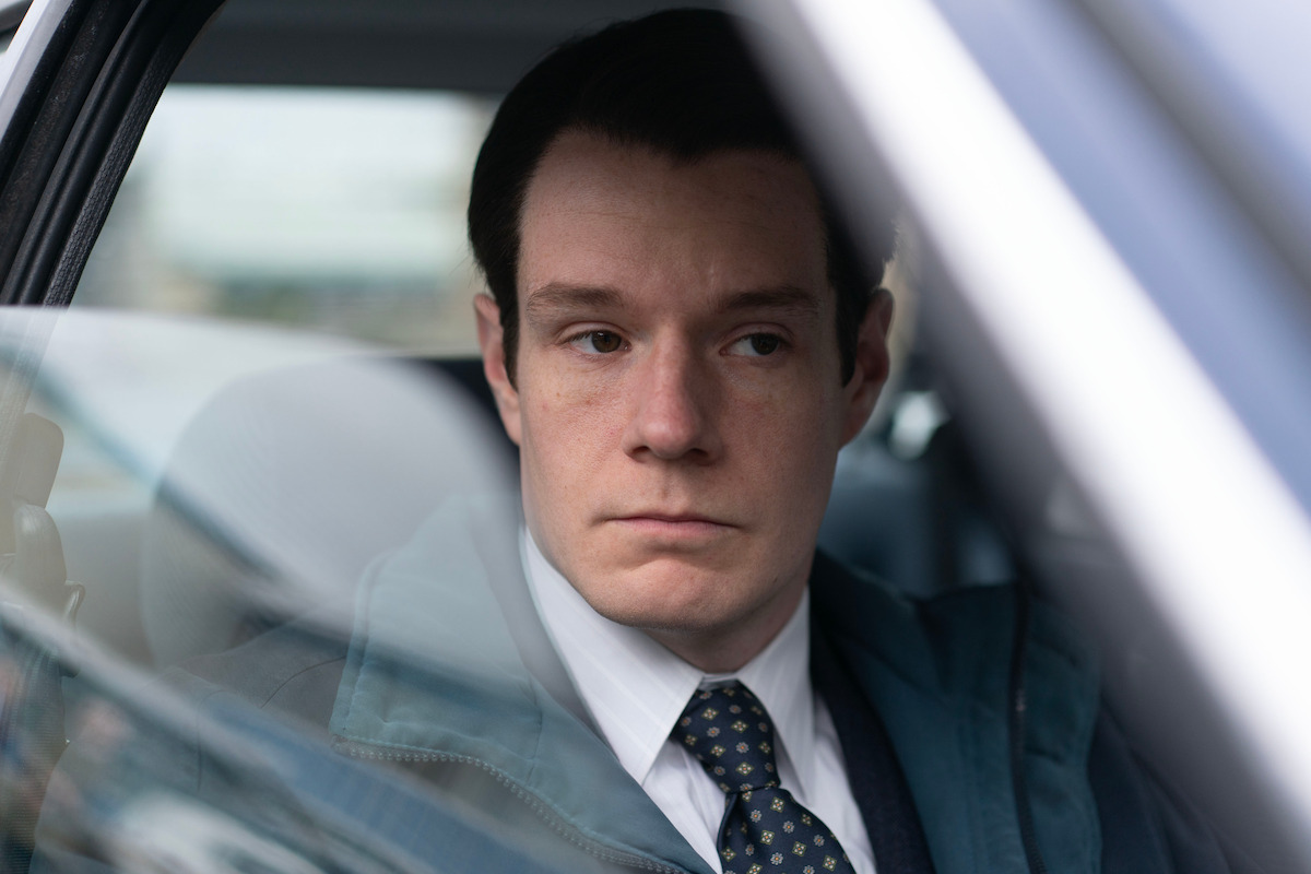 Man in a suit and tie sitting in a car, looking serious and gazing out of the window. The setting appears to be urban, seen through the car window, suggesting a thoughtful or tense moment.