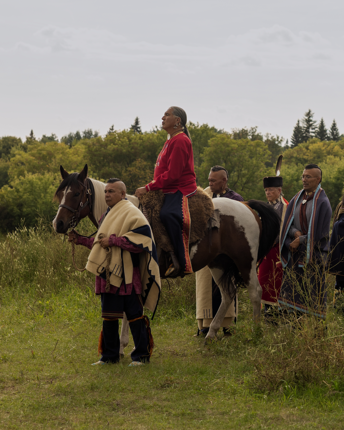 A group of Indigenous people in traditional clothing stand in a grassy field with trees in the background; one person sits on a horse while others walk beside, suggesting a cultural or ceremonial gathering outdoors.