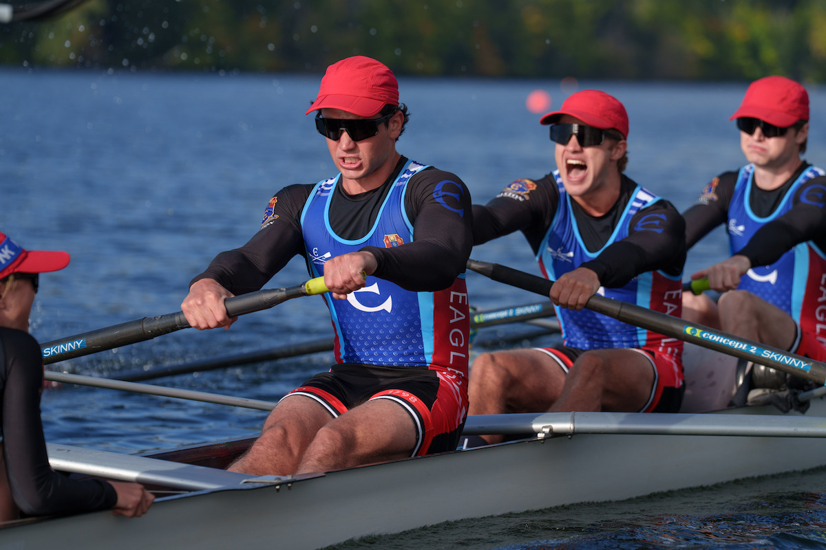 Rowing team of four men in matching uniforms and red caps competing on a lake, focused and exerting effort as they row together during a race with green trees and buoys visible in the background.