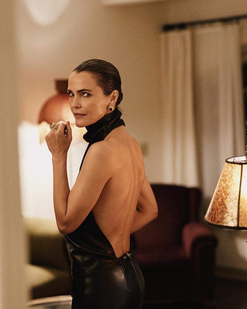Woman in a backless black dress stands indoors, looking over her shoulder in a warmly-lit living room with a lamp, armchair, and curtains in the background.