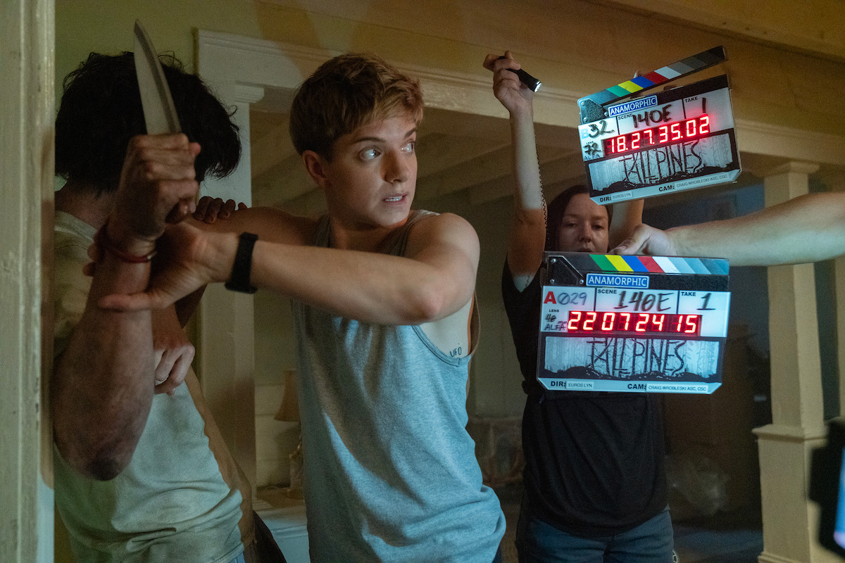 A movie scene being filmed indoors, featuring a young actor grabbing an attacker’s arm holding a knife, with two crew members using clapperboards, indicating a tense action setting on a film set.