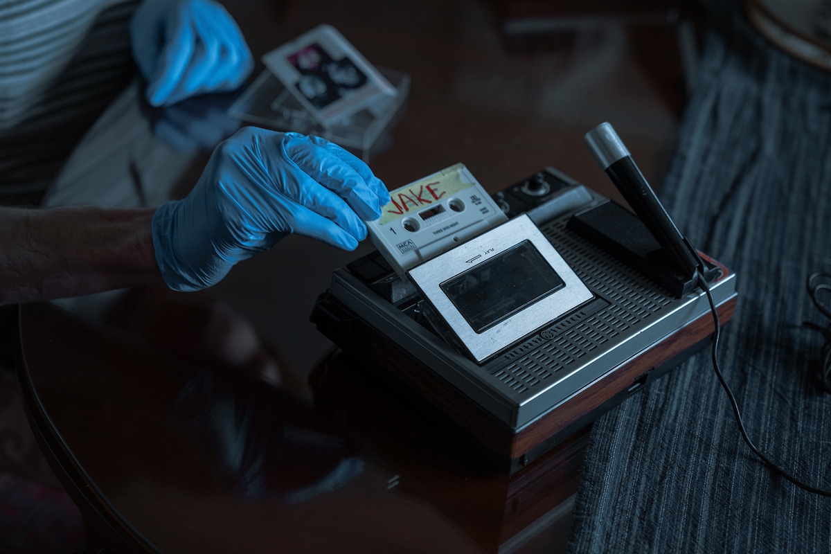 Person wearing blue gloves inserts a labeled cassette tape into a vintage tape recorder with a microphone on a wooden table, suggesting an investigative or archival environment.