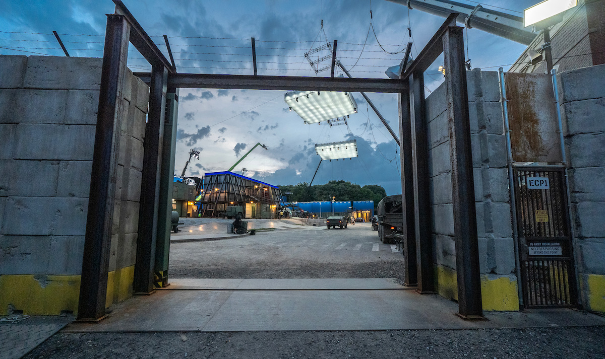 Camera looks through a tall, unfinished gate onto a movie set styled as a secure military compound at dusk.