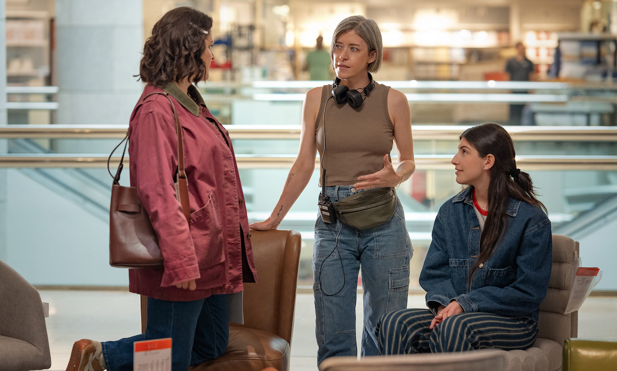Three women talking in a modern indoor space resembling a store or mall, with two seated on display chairs and one standing, engaged in conversation among furniture and retail displays.