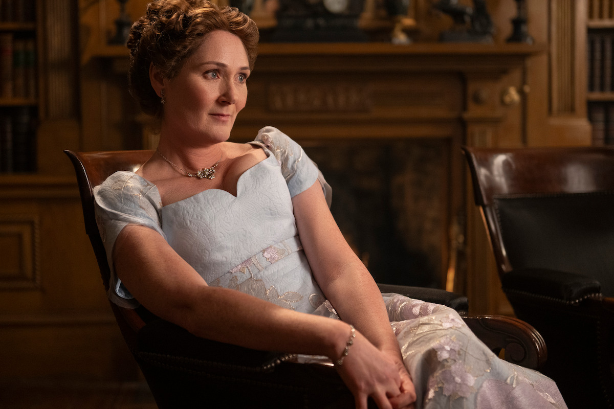 A woman in an elegant period dress sits in a wooden chair in a richly decorated room with bookshelves and a fireplace, appearing calm and contemplative. The atmosphere is refined and historical.