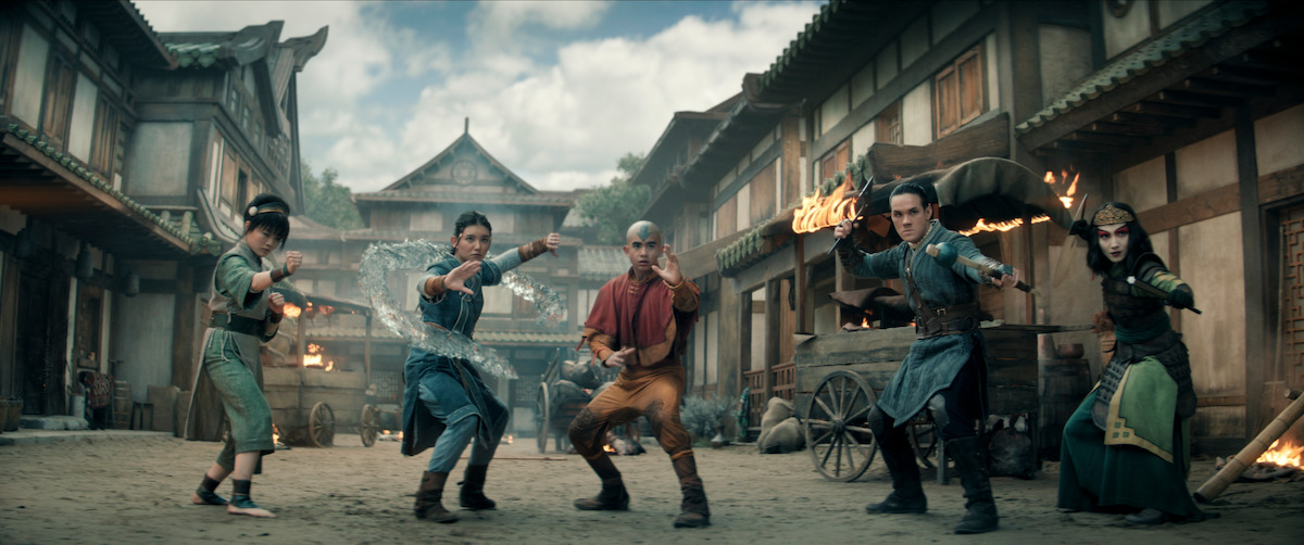 Toph, Katara, Aang, Sokka, and Suki in martial arts stances, ready for battle in a historic village street, surrounded by wooden buildings with smoke and fire in the background, suggesting an action scene with fantasy elements.