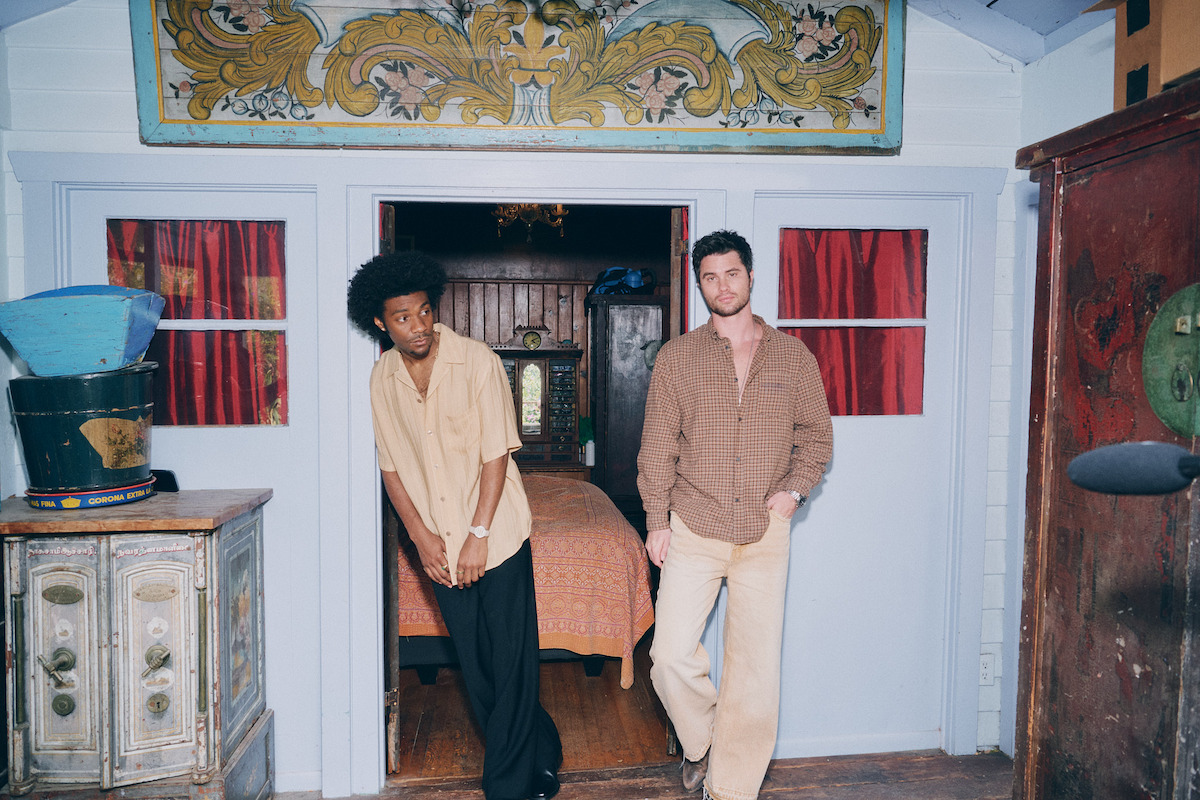 Two men standing in front of a rustic, ornately decorated room with blue wooden walls and red curtains, one leaning against the doorframe, vintage furniture and decorative elements visible around them.