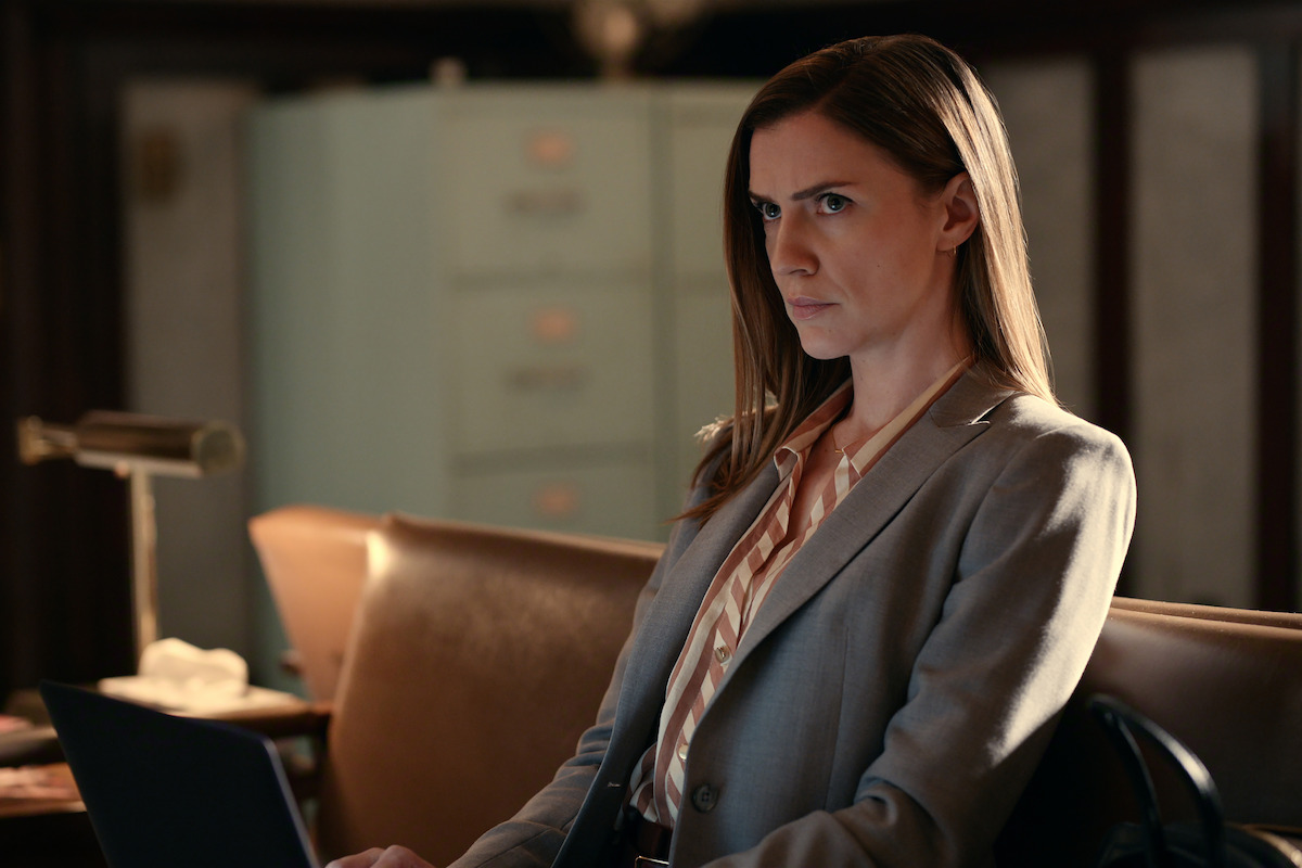 A woman in a business suit sits on a brown couch, working on a laptop in an office with a vintage lamp and filing cabinets, looking serious and focused.