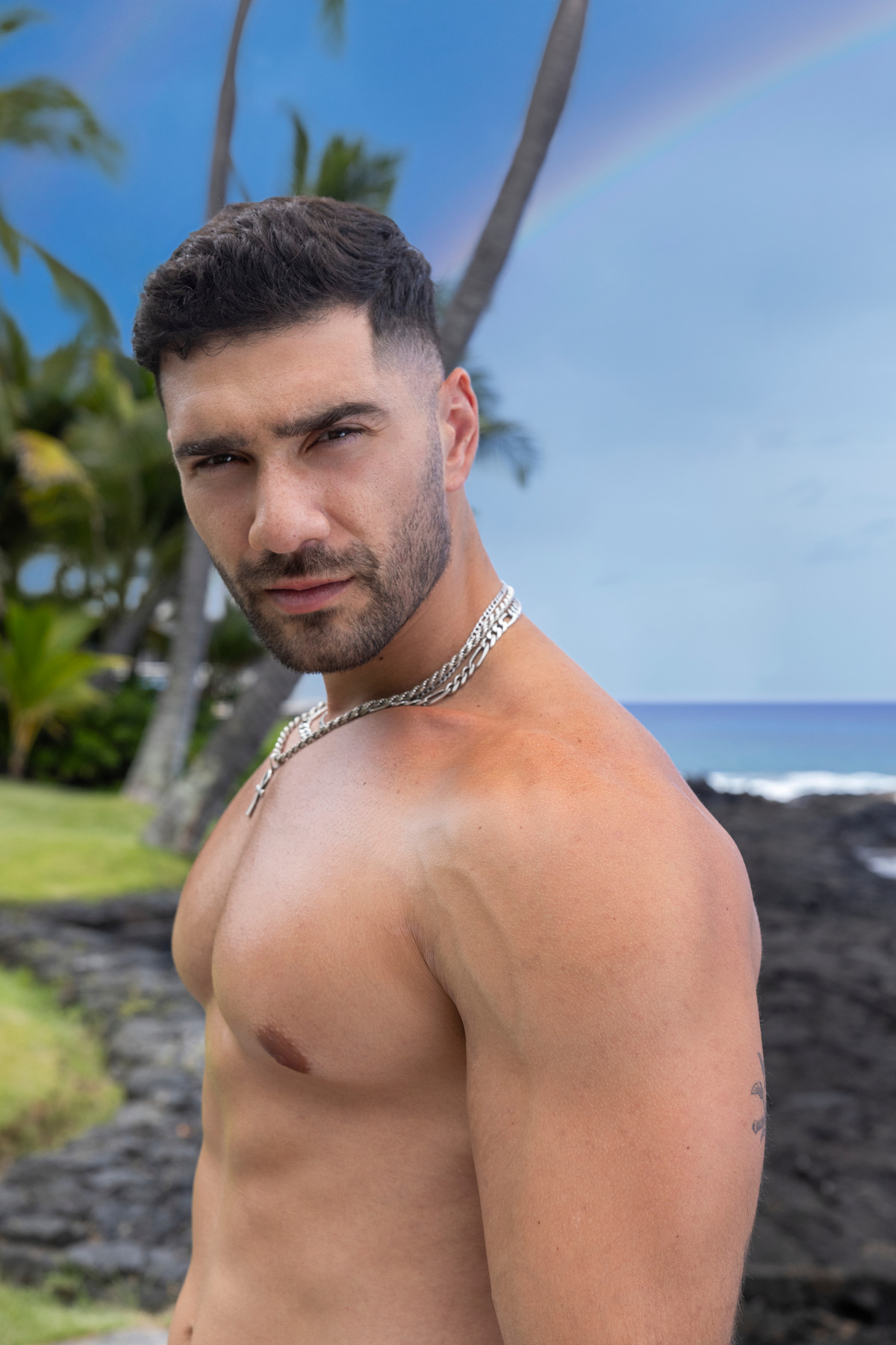 Shirtless man with short dark hair and chain necklace standing on a tropical beach with black rocks, palm trees, blue sky, and a rainbow in the background.