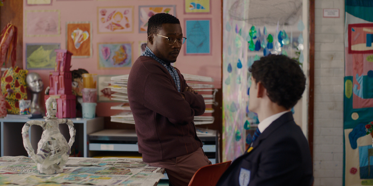 Two people talking in a colorful art classroom with student artwork on the walls, art supplies, and ceramic sculptures on the table, creating a creative, educational environment.
