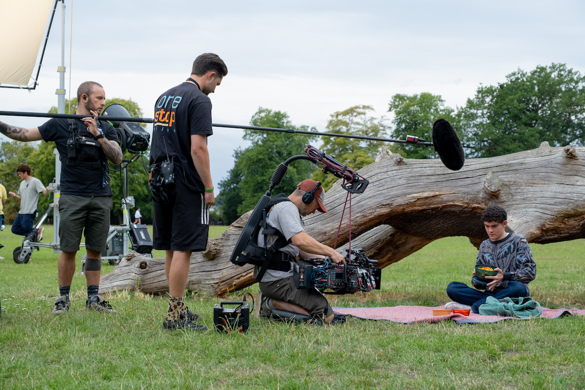 Film crew with camera and boom mic record a young actor seated on a blanket next to a fallen tree in an outdoor park setting, with equipment and crew members visible, under cloudy skies.