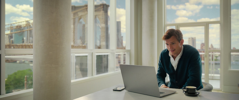 Ashton Kutcher sits in front of computer in front of windows looking out onto the Brooklyn Bridge  in Your Place or Mine.