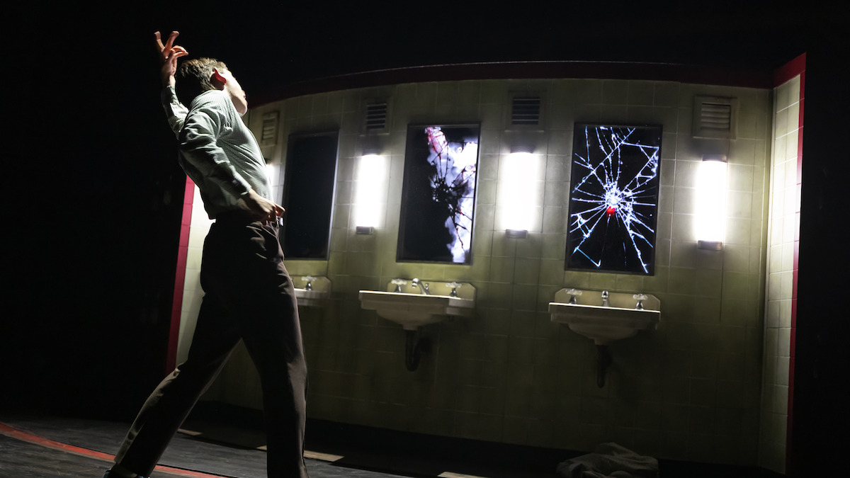 A person dramatically poses in a dimly lit public restroom set with broken mirrors and sinks, creating a tense and emotional atmosphere.