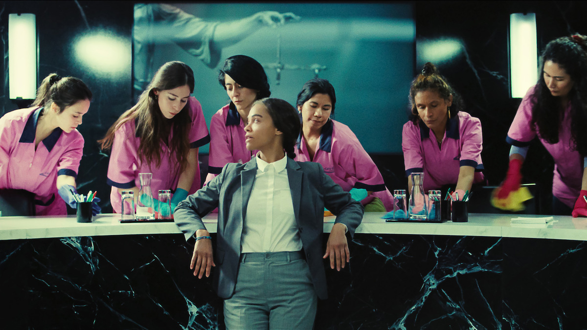 Rita (Zoe Saldaña) wears a grey suit and white shirt and a bunch of people wearing pink outfits stand around her.