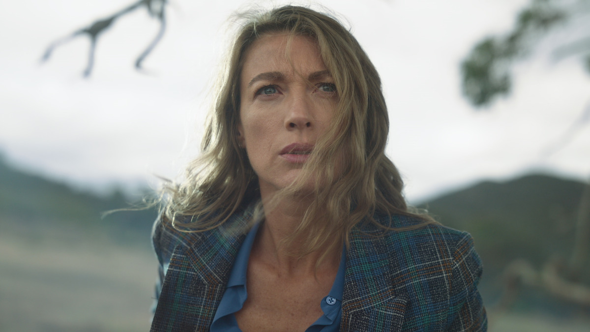 A concerned woman with long blonde hair wearing a blue plaid blazer outdoors, with blurred mountains and branches in the background, suggesting a dramatic or tense moment in a natural setting.