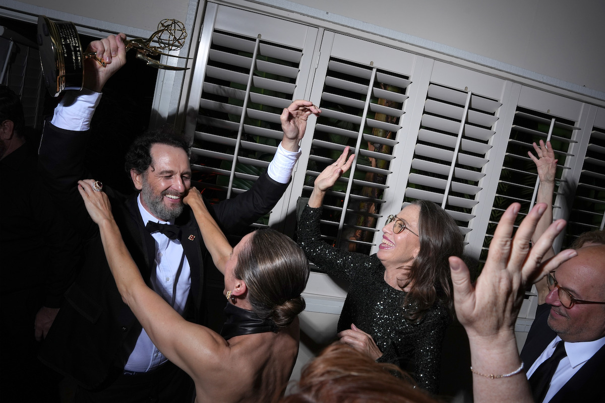 A man in a tuxedo celebrates with friends indoors, holding up an award trophy. People surround him, smiling and reaching out in excitement, suggesting a festive, joyful atmosphere at an awards event or party.
