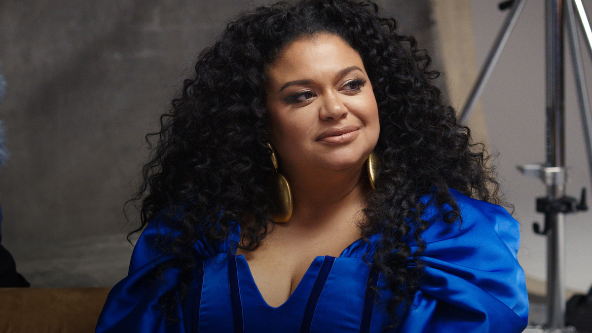 Woman with long curly hair wearing a vibrant blue dress and large gold earrings, seated indoors against a gray backdrop with studio lighting equipment visible in the background.