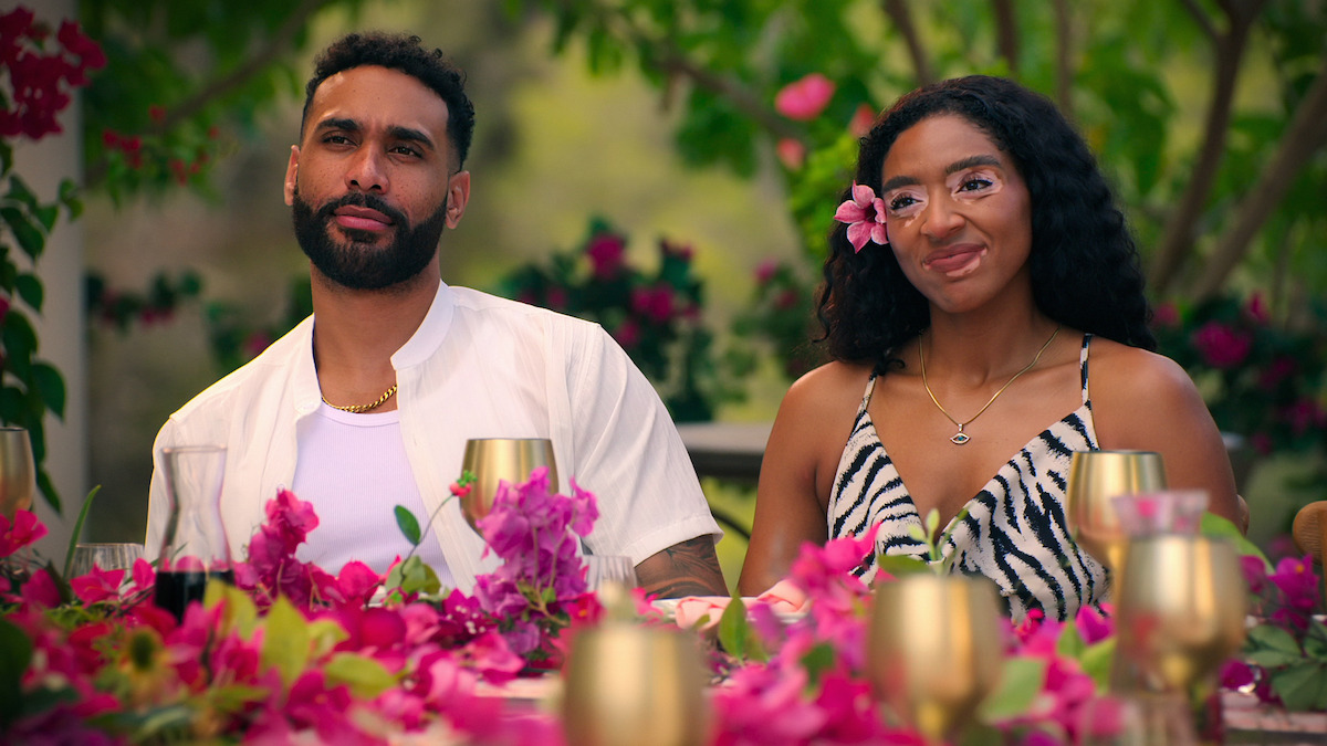 Two people sit at a floral outdoor table, surrounded by vibrant flowers and golden cups, with greenery in the background. The woman has a flower in her hair and both are dressed in summer attire, suggesting a festive, relaxed gathering.