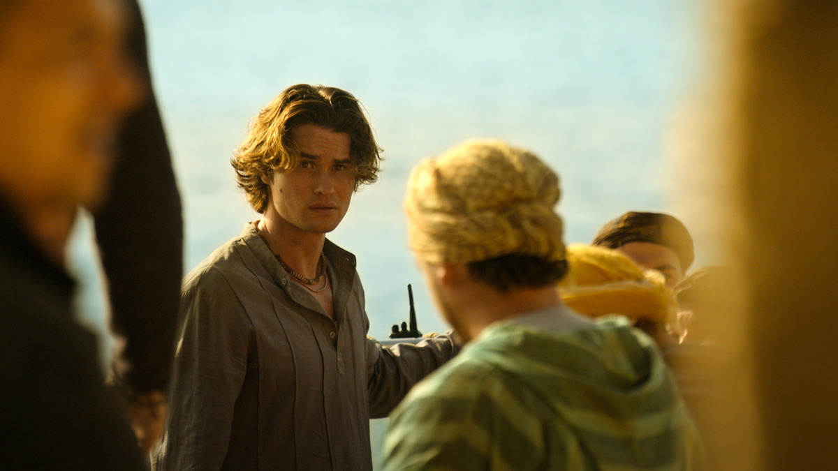 A young man with wavy hair stands outdoors near water, holding a walkie-talkie, looking toward the camera, surrounded by people dressed in casual clothing under warm sunlight.