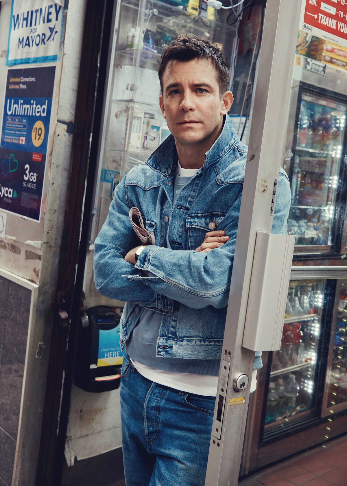 Man in denim jacket and jeans stands in the doorway of a convenience store, arms crossed, with various product displays and posters visible inside the shop. Urban setting with a casual atmosphere.