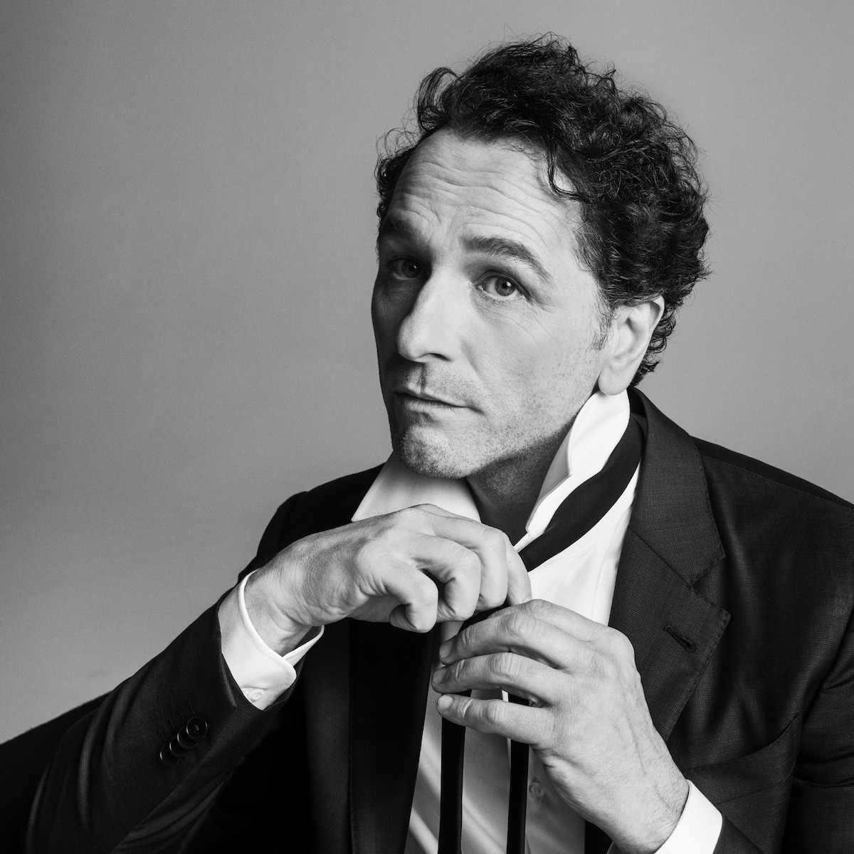 Man in a suit adjusting his tie in a studio with a neutral background, black-and-white photo, contemplative expression, sophisticated and introspective mood.
