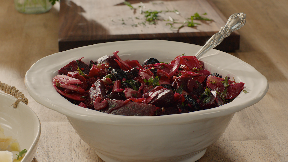 A white bowl filled with beet salad, garnished with herbs, sits on a wooden table with a vintage spoon. The environment is bright and rustic, featuring a cutting board with fresh herbs in the background, evoking a cozy, homemade feel.