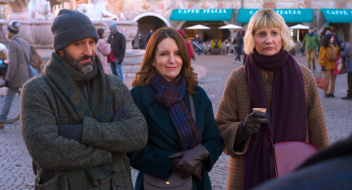 Three adults in winter clothing stand together in a European outdoor plaza, with a cafe and people in the background. They appear to be having a casual conversation on a chilly day.