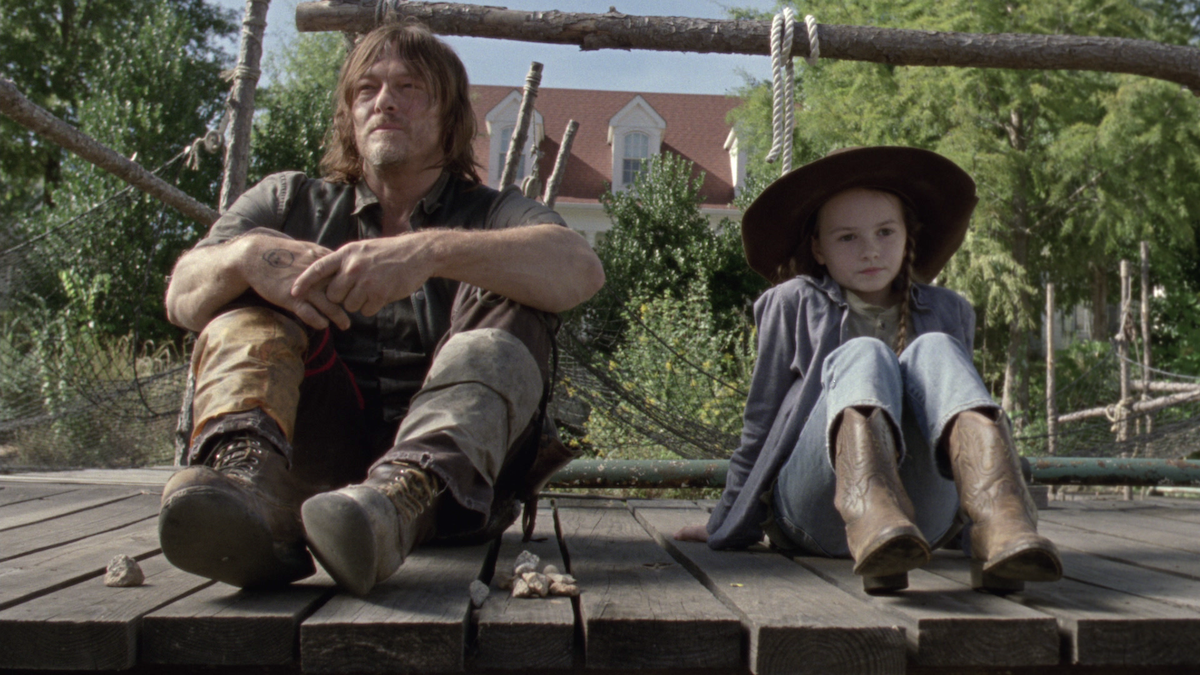 Man and young girl sit side by side on a wooden platform outdoors, surrounded by greenery and rustic wooden railings, with a large house in the background on a sunny day.