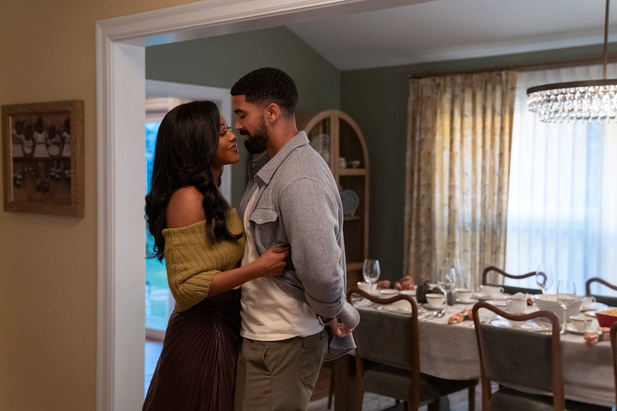 A couple stands close together in a warm, cozy dining room, smiling at each other. The table behind them is set for a meal, with plates, glasses, and silverware, and sunlight streams through sheer curtains on the window.