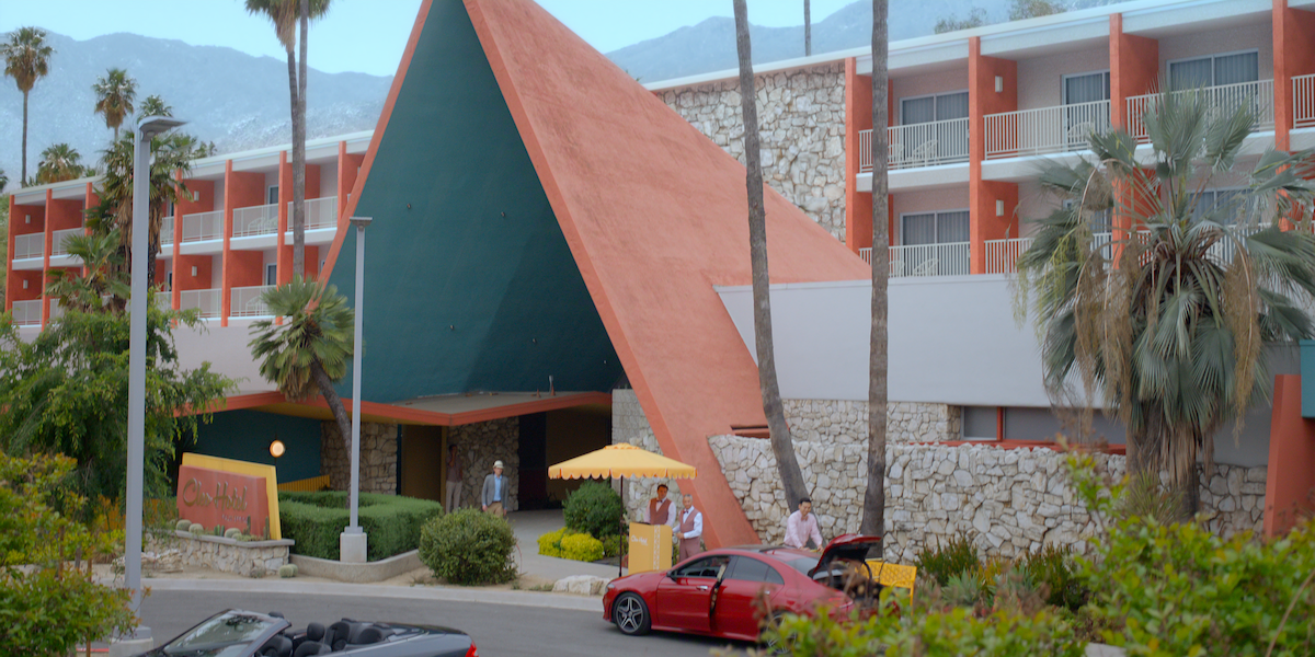 A retro-style hotel with a triangular entrance and stone walls, surrounded by palm trees and mountains. People stand by a red car with its trunk open at the front driveway.