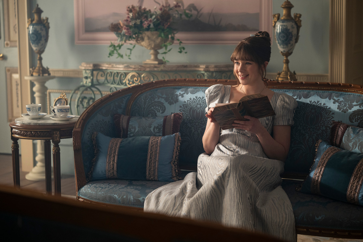 A woman in a Regency-era dress sits on an ornate blue sofa, smiling while reading a book. The elegant room features antique decor, a tea set on a side table, vases, and a painting, evoking a historical, refined atmosphere.